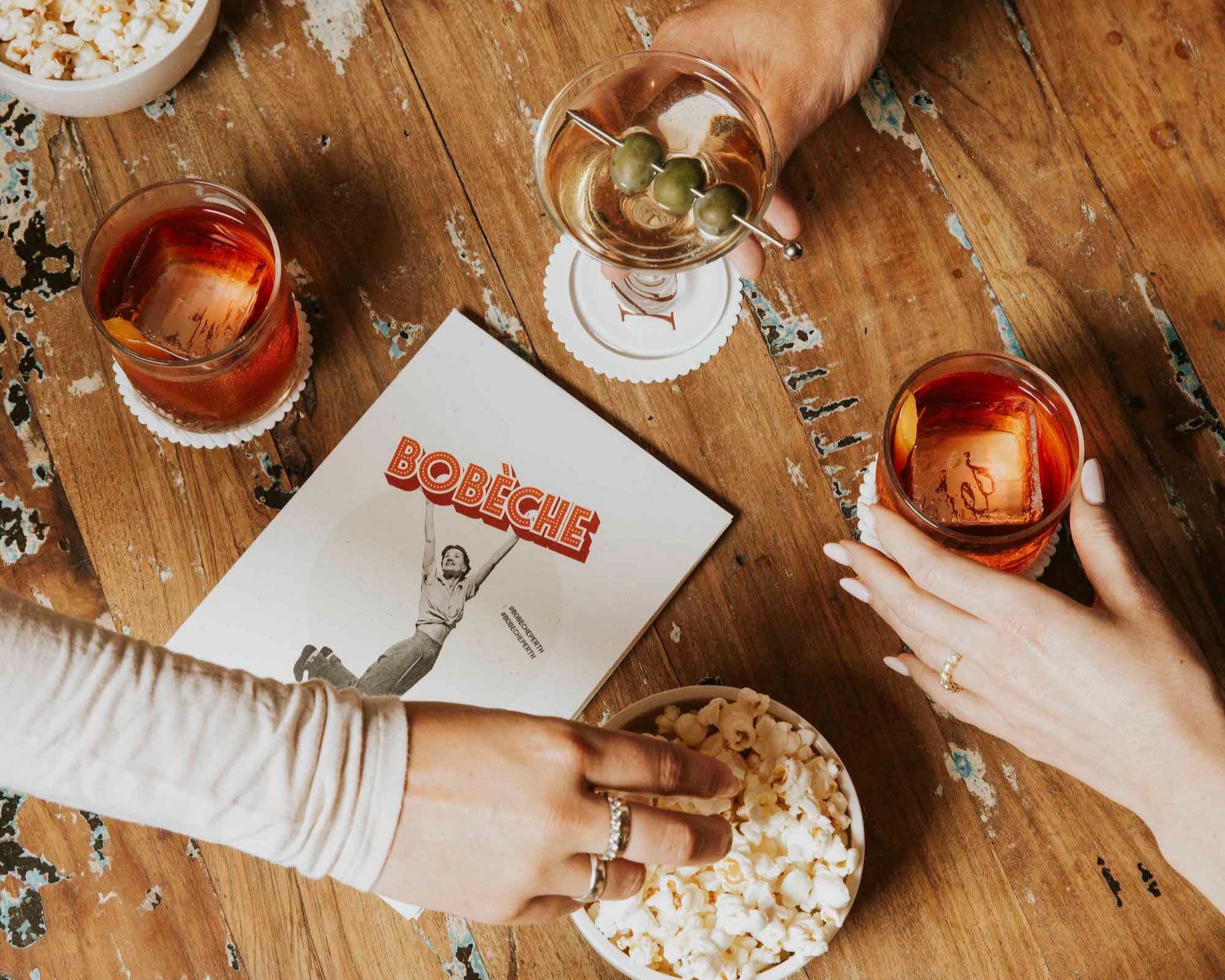 Two people enjoying drinks and popcorn on a wooden table with a magazine titled 'Bobbette'.